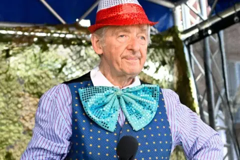Uśmiechnięty Andrzej Rosiewicz w cekinowym kapeluszu i muszce, trzymający mikrofon. Wiek artysty to 80 lat.