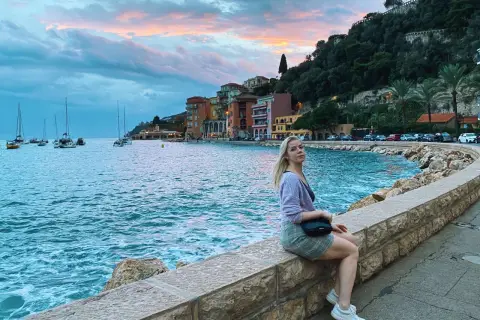Kobieta siedzi na kamiennym murku nad morzem w Villefranche-sur-Mer, podziwiając zachód słońca i żaglówki.
