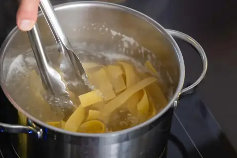 Szef kuchni dodaje szczypcami szeroki makaron pełnoziarnisty do gotującej się wody w garnku.