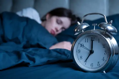 Frau schläft tief, während ein Wecker die Zeit anzeigt. Wie viel Schlaf braucht man mit 40? Die Antwort liegt im erholsamen Schlaf.