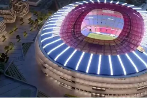 Nowe Camp Nou w nocnej odsłonie, z nowoczesną, oświetloną fasadą i dachem. Widać fragmenty otoczenia z budynkami i zielenią.