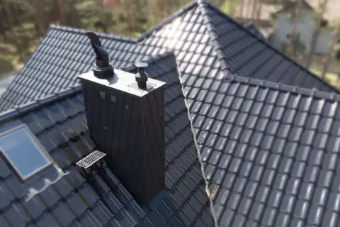 Nowoczesny, czarny komin stalowy na błyszczącym, ciemnym dachu. W tle drzewa.