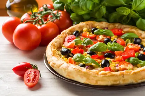 Domowa pizza z pomidorami, oliwkami i kukurydzą, gotowa do pieca. Obok świeże pomidory i bazylia.