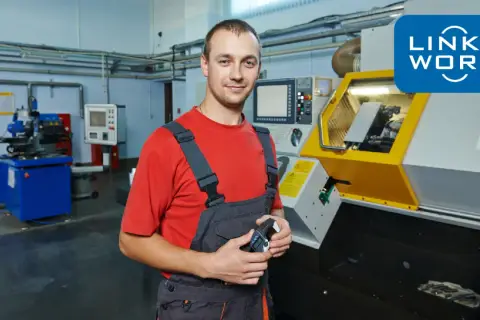 Uśmiechnięty tokarz w warsztacie, trzymający przyrząd pomiarowy, obok nowoczesnej maszyny CNC.