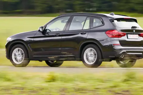 Ciemnoszary BMW G01 pędzi po wiejskiej drodze, rozmazując zielone pola i drzewa w tle.