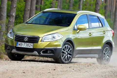 Zielony Suzuki S-Cross na drodze leśnej, wzbudzający pozytywne suzuki s-cross opinie. Kurz spod kół świadczy o jego terenowych możliwościach.