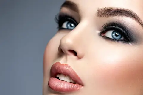 Niebieskie oczy z mocnym makijażem typu smokey eyes, podkreślone długimi rzęsami i ciemnymi cieniami. Usta w odcieniu nude.