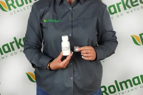 Osoba w szarej koszuli z logo Rolmarket trzyma buteleczkę z płynem i miarkę. To może być środek na chwasty, który pomoże w walce z niechcianą roślinnością.