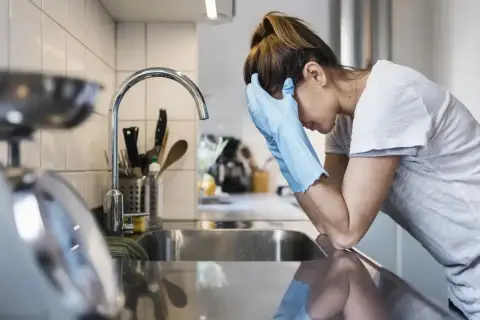 Kobieta w rękawiczkach opiera się o zlew, wyraźnie przygnębiona. Mizofobia utrudnia jej codzienne czynności.