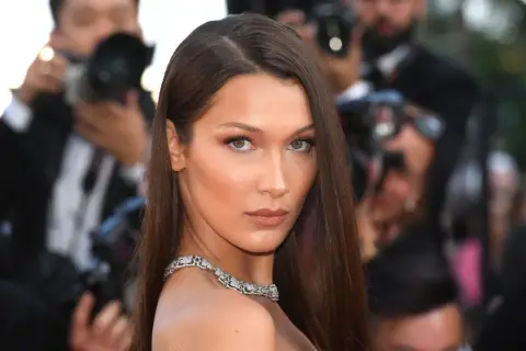 Bella Hadid con un elegante side part, luciendo un collar de diamantes y rodeada de fotógrafos.