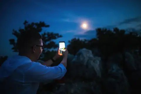Chłopak z telefonem Samsung próbuje zrobić zdjęcie księżyca. Nocne niebo, drzewa i skały tworzą tło.