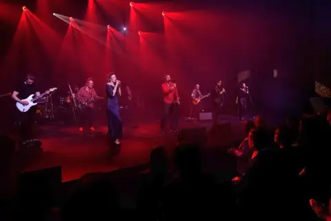 Zespół muzyczny na scenie, oświetlony czerwonymi światłami. Wokalistka w granatowej sukni, obok niej gitarzyści i wokalista w czerwonej koszuli. W tle widać perkusję i innych muzyków. Publiczność siedzi w ciemności. To jakby **sufler** podpowiadał im k...