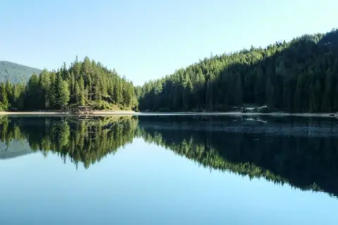 Spokojna tafla jeziora odbija gęsty las i góry. Widok na malowniczą kotlinę z drewnianym pomostem.