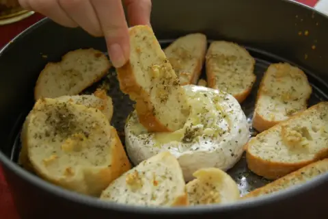 Ciepły serek camembert w okrągłej formie, otoczony kromkami pieczywa z ziołami. Ktoś zanurza kawałek chleba w rozpływającym się serze.