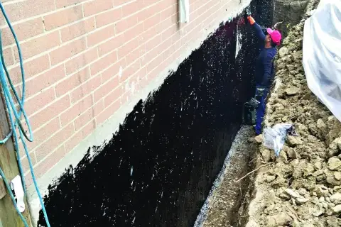 Pracownik zabezpiecza ławy fundamentowe czarną masą izolacyjną.