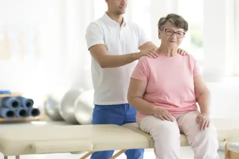 Starsza pani na rehabilitacji. Fizjoterapeuta wspiera ją podczas ćwiczeń w sanatorium ortopedycznym NFZ.
