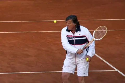 Panatta w białej kurtce i spodenkach, z rakietą i piłką tenisową na korcie.