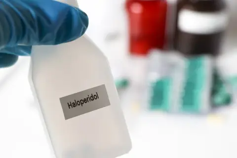 Blauer Handschuh hält Flasche mit der Aufschrift "Haloperidol". Im Hintergrund sind Medikamentenpackungen zu sehen.