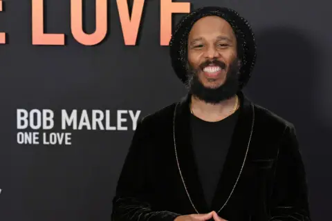 Uśmiechnięty Ziggy Marley na tle napisu "Bob Marley: One Love". Wiele dzieci Boba Marleya kontynuuje jego dziedzictwo.