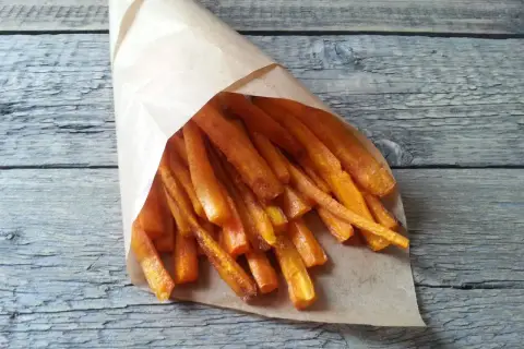 Chrupiące frytki z marchewki w papierowym rożku, ułożone na drewnianym blacie. Zdrowa przekąska.