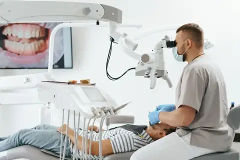 Stomatolog w masce i rękawiczkach bada pacjenta pod mikroskopem. Na ekranie widać zbliżenie zębów.