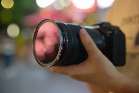 Dłoń trzyma aparat z obiektywem, gotowy do uchwycenia chwili. Czerwony pierścień obiektywu i fioletowy element przy **shutterze** dodają mu charakteru.