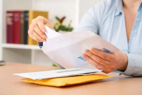 Dłoń wkłada białą kartkę do koperty. Czy można wysłać list bez nadawcy? Na biurku leżą koperty.