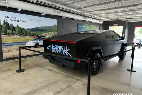Czarny Tesla Cybertruck z neonowym napisem "CYBERTRUCK" na tyle. W tle reklama "Electric All-Wheel Drive". Cena tego futurystycznego pojazdu budzi zainteresowanie.