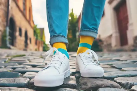 Kolorowe skarpetki do sneakersów dodają charakteru stylizacji. Osoba w jeansach i białych butach idzie po bruku.