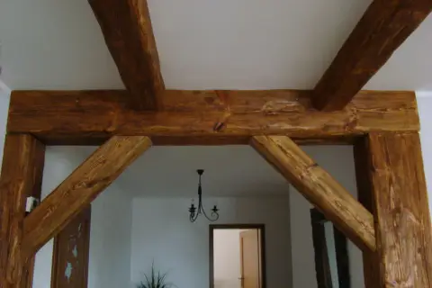 Solidne łączenie belek drewnianych pod kątem prostym, tworzące solidną konstrukcję. W tle widać drzwi i żyrandol.