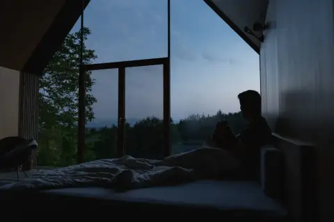 Mann sitzt im Bett, blickt aus dem Fenster in die Dämmerung. Ein ruhiger Moment, der den schlaf wach rhythmus widerspiegelt.