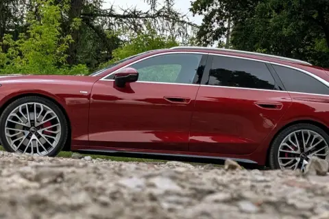 Lśniące, czerwone Audi A6 C9 Avant stoi na kamienistym podłożu, otoczone zielenią.