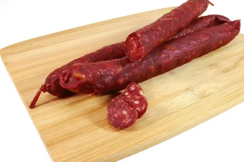 Trzy długie kiełbaski chorizo leżą na drewnianej desce. Jedna jest przekrojona, ukazując charakterystyczny, czerwony środek.
