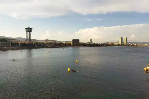 Vista de la playa y el mar con el icónico hotel vela al fondo. Personas disfrutan del agua en tablas de paddle surf.