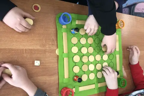 Dzieci grają w grę planszową, która jest częścią zajęć korekcyjno kompensacyjnych. Na zielonej macie znajdują się drewniane pionki i kostka.