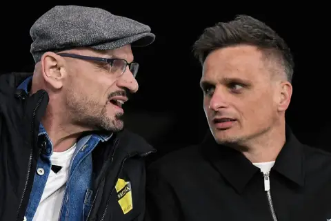 Dwóch mężczyzn rozmawia. Jeden w czapce i okularach, drugi z charakterystyczną fryzurą. Na kurtce jednego z nich widnieje napis "Wieczysta Kraków".