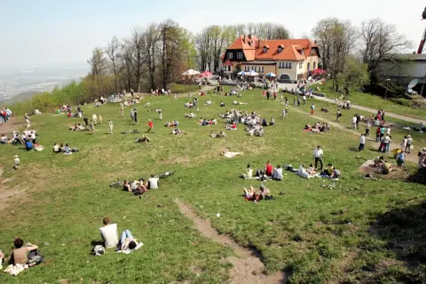 Ludzie odpoczywają na trawie na zboczu góry. W tle budynek z czerwonym dachem. Widać ścieżki i drzewa.