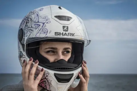 Dziewczyna w białym, ozdobnym kasku na skuter marki Shark. Jej oczy patrzą w bok, a dłonie poprawiają zapięcie.
