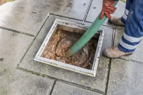 Szambonurek w rękawicach i niebieskich spodniach z odblaskami używa zielonego węża do opróżniania szamba.