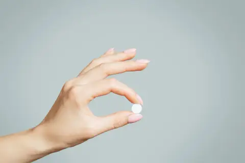 Dłoń z różowymi paznokciami trzyma białą tabletkę, być może zolpidem, na jasnoszarym tle.