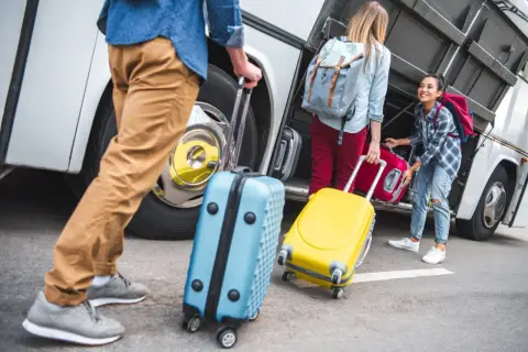 Ludzie z walizkami przy autobusie. Jedna walizka jest niebieska, druga żółta. W tle widać bagażnik autobusu.