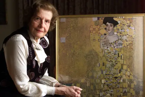 An elderly woman poses beside a framed woman in gold painting, her expression serene.