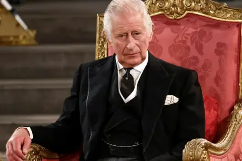 Król Karol w wieku siedemdziesięciu lat, w eleganckim czarnym garniturze, siedzi na ozdobnym tronie.