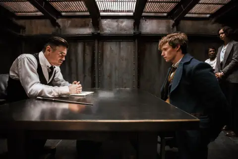 Scena z filmu "Fantastyczne zwierzęta i jak je znaleźć". Newt Scamander i Percival Graves siedzą przy stole, podczas gdy dwie kobiety stoją w tle.