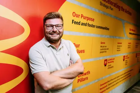 Uśmiechnięty mężczyzna w okularach na tle ściany z hasłami McDonald's. Zastanawiasz się, ile jest McDonaldów w Polsce? Ta ściana może pomóc w rozwoju sieci.