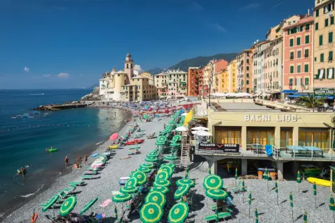 Plage animée à Camogli, Ligurie, avec des parasols verts et jaunes, des bâtiments colorés et la mer bleue.