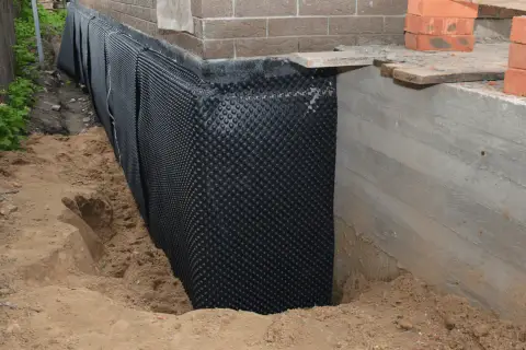 Folia kubełkowa chroni betonowy cokół przed wilgocią. Widać wykop i materiały budowlane.