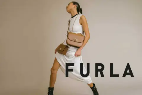 Modelka w białej sukience prezentuje dwie brązowe torebki Furla, symbol tanich włoskich marek.