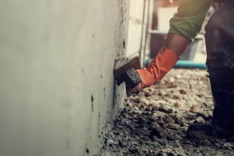 Pracownik w pomarańczowych rękawicach tynkuje ścianę. Brak zgody sąsiada na ocieplenie budynku może opóźnić prace.