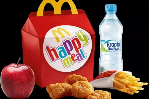 Czerwone pudełko z napisem "happy meal", obok jabłko, nuggetsy, frytki i butelka wody. Taka hepi mil cena to świetna opcja dla dzieci.